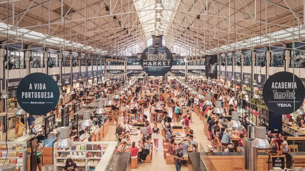Belebter Time Out Market in Lissabon. Menschen sitzen an langen Tischen, essen und unterhalten sich. Überall gibt es Essensstände.