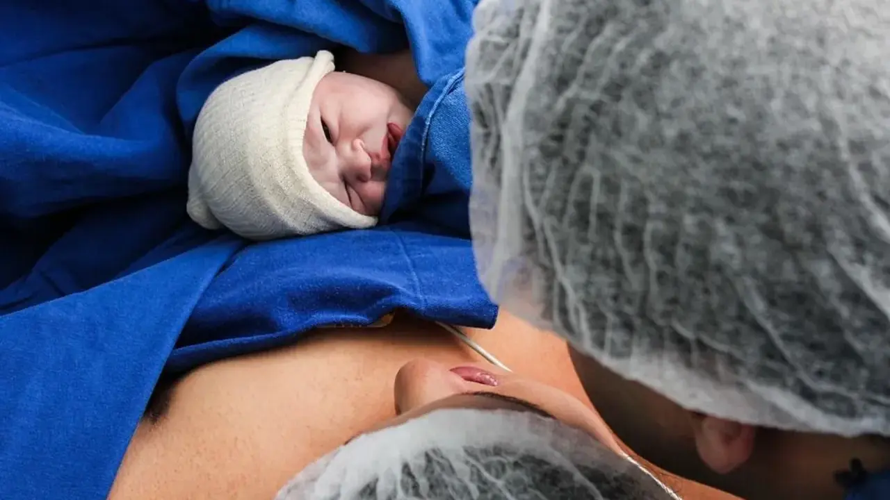 Noworodek w czepku, otulony niebieskim materiałem, leży na brzuchu mamy. Widoczna twarz dziecka i fragment twarzy mamy. To piękny moment porodu lotosowego.