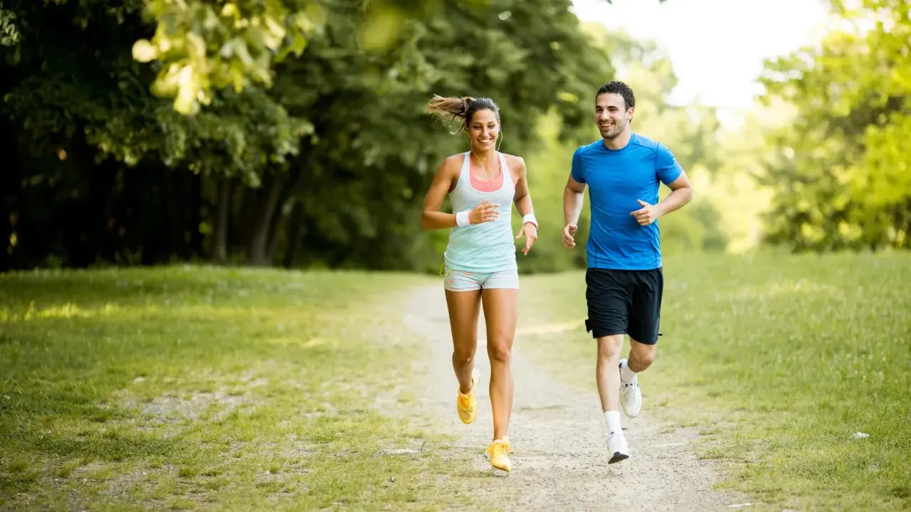 Ein lachendes Paar joggt glücklich durch einen sonnigen Park. Sie genießen die frische Luft und die Bewegung – das ist, was Joggen bringt.