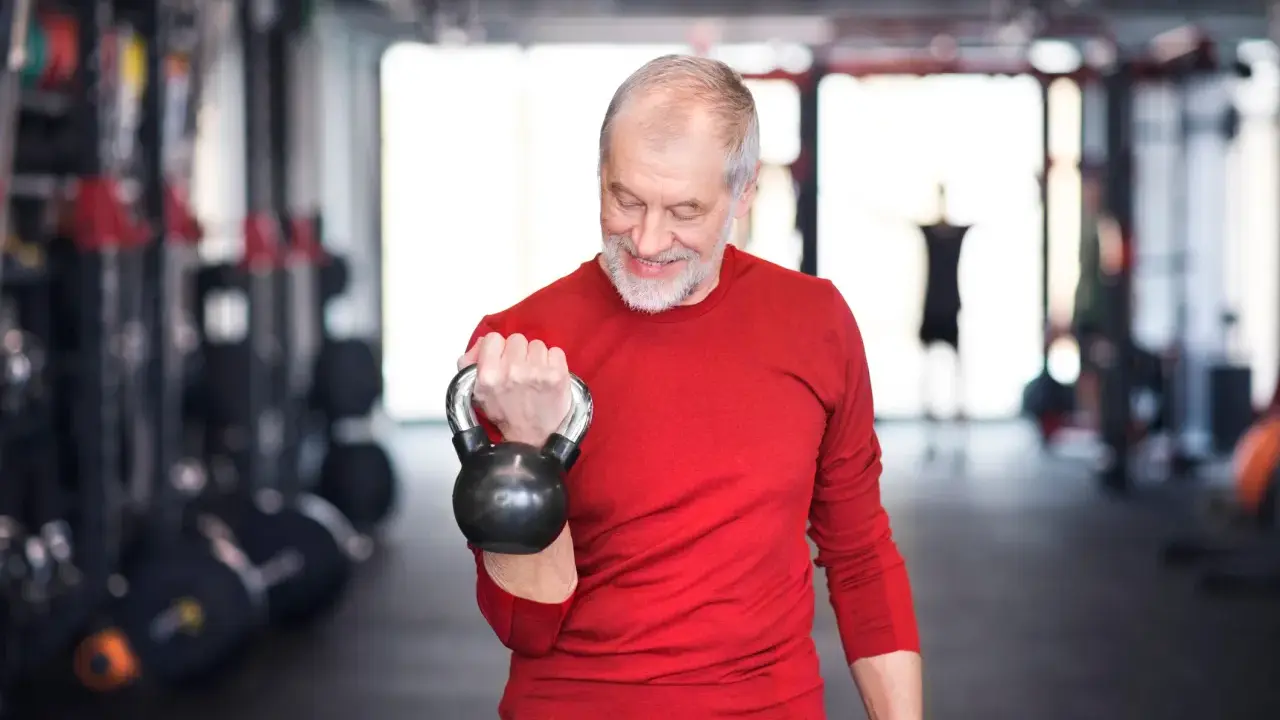 Ein älterer Mann mit Bart trainiert im Fitnessstudio mit einer Kettlebell. Ein Beispiel für Krafttraining ab 60 Jahren Trainingsplan.