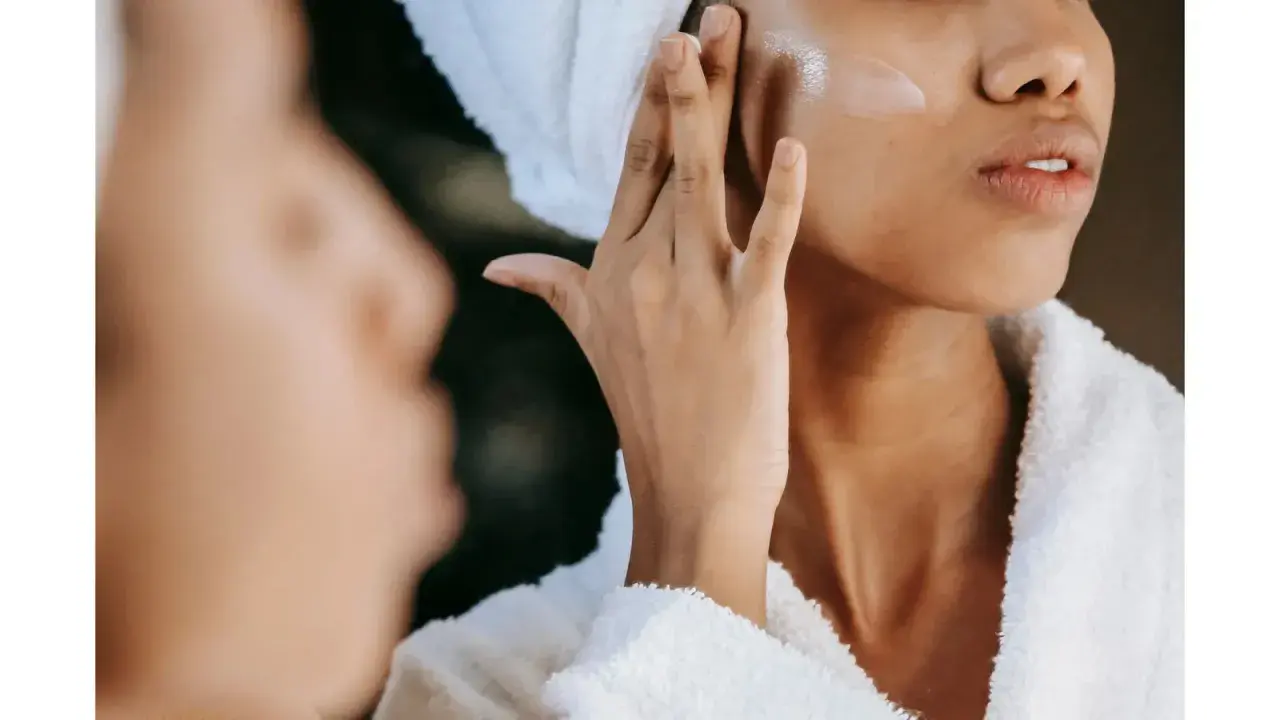 Une femme applique une crème pour l'hydratation du visage, enveloppée dans une serviette et un peignoir douillet.