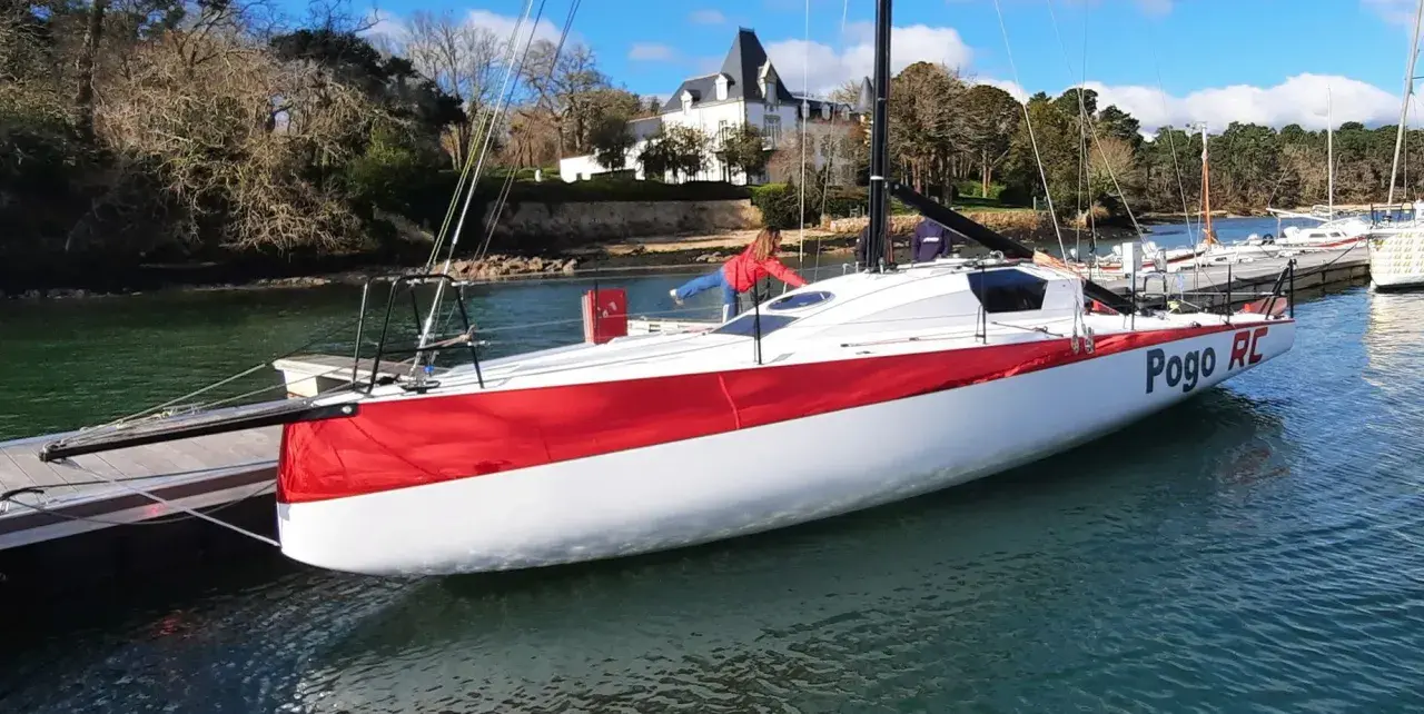 Le voilier Pogo RC, blanc et rouge, est amarré à un ponton. Une personne en blouson rouge est à bord.