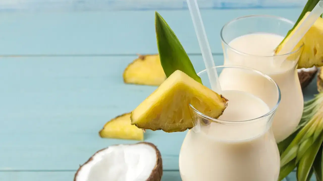 Dwa orzeźwiające koktajle Pina Colada, ozdobione kawałkami ananasa i liśćmi, na tle niebieskich desek. Idealny przepis na lato!