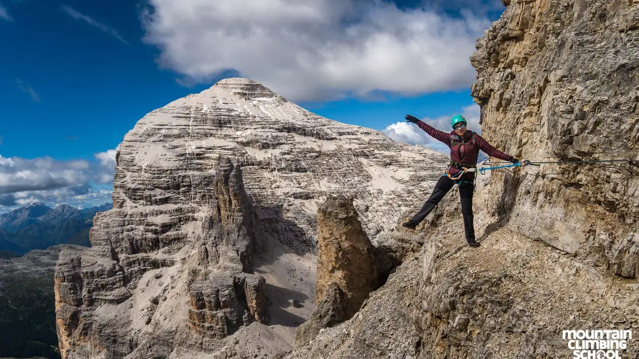 Kobieta na via ferrata dla początkujących, z rękami rozłożonymi na boki, na tle skalistych gór i błękitnego nieba.