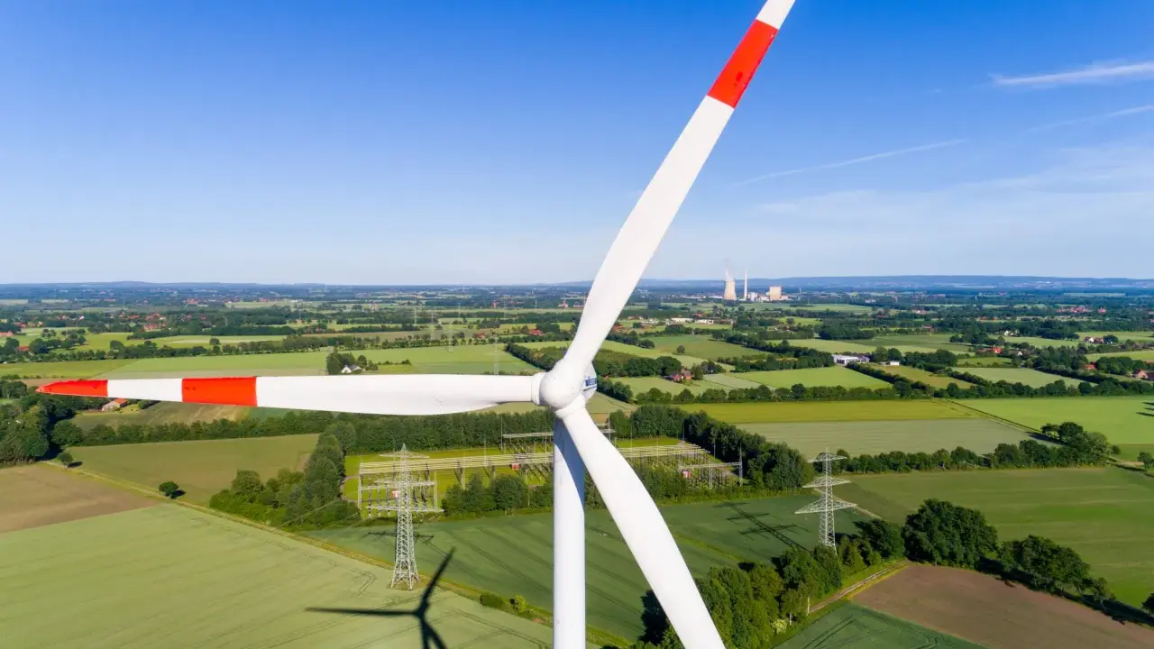 Windkraft Energie: Ein riesiger Windpark dominiert die Landschaft, umgeben von grünen Feldern und Bäumen. Im Hintergrund sind Kühltürme zu sehen.