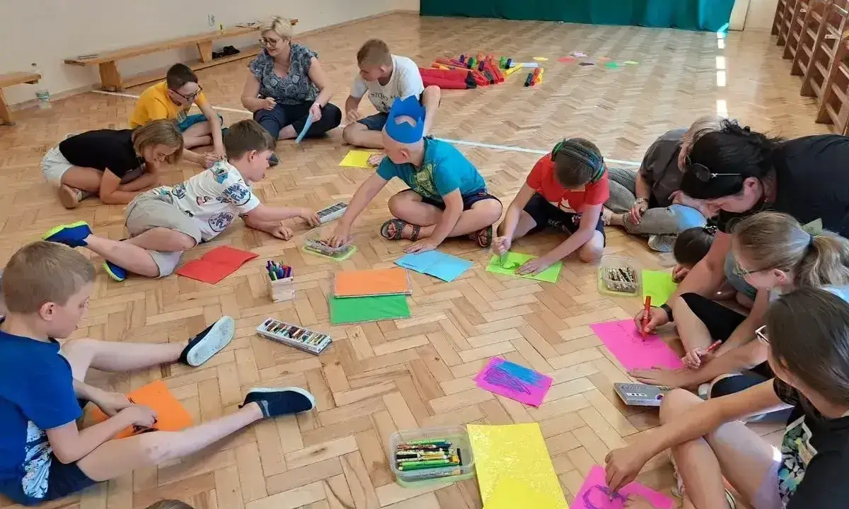 Dzieci i dorośli siedzą na podłodze, tworząc kolorowe rysunki. Wszyscy biorą udział w wesołych zabawach integracyjnych, rozwijając kreatywność.