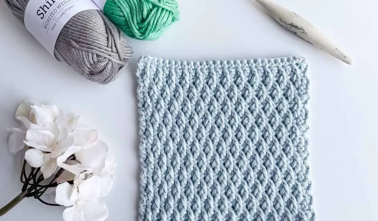 Muestra de tejido a crochet con el patrón nudo de salomon en color azul claro, junto a ovillos de lana gris y verde, una flor blanca y un ganchillo.
