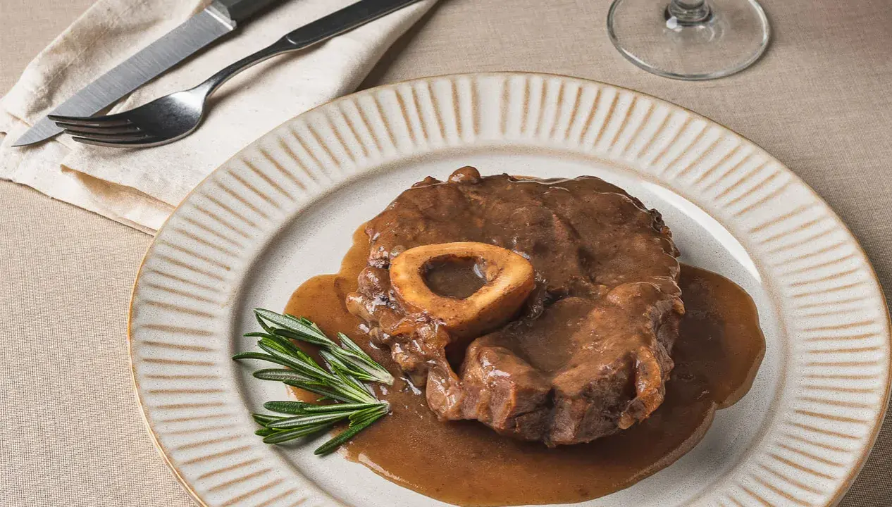 Un delicioso osso buco, tierno y jugoso, bañado en su salsa, servido en un plato con romero fresco.