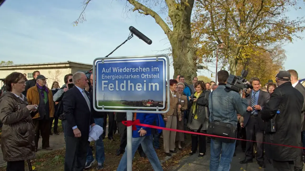 Feldheim, ein Vorreiter unter den energieautarken Gemeinden Deutschlands, wird hier mit einem Schild gefeiert. Eine Gruppe von Menschen, darunter ein Kameramann, versammelt sich.