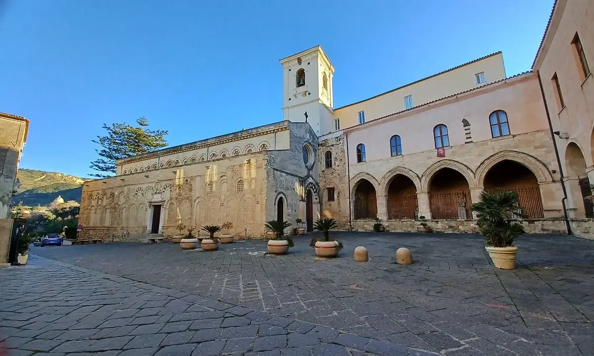 La piazza antistante la chiesa di Tropea, con il suo campanile e gli archi del chiostro, sotto un cielo azzurro.