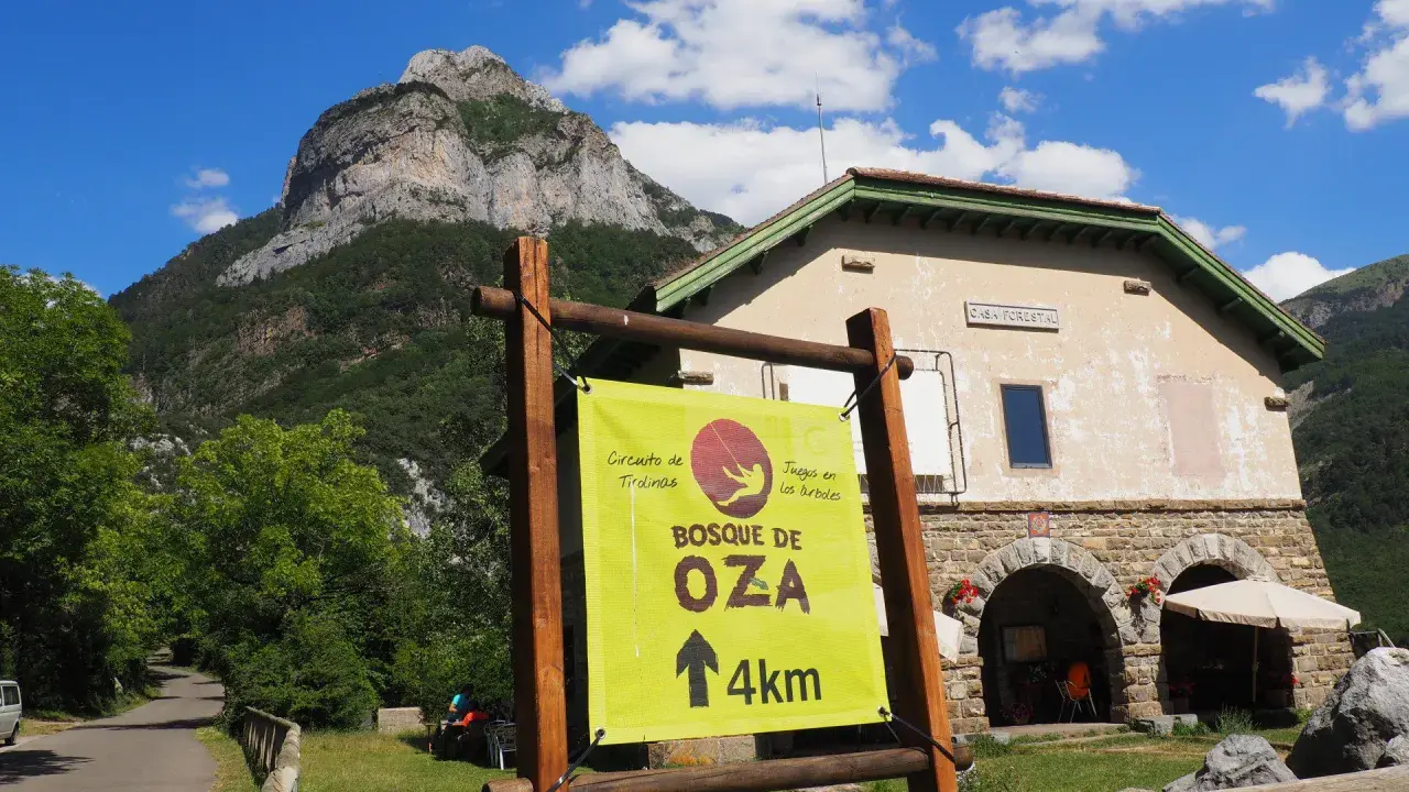 Cartel indica "Bosque de Oza 4km" en un día soleado en el valle de hecho, con una casa forestal y montañas al fondo.