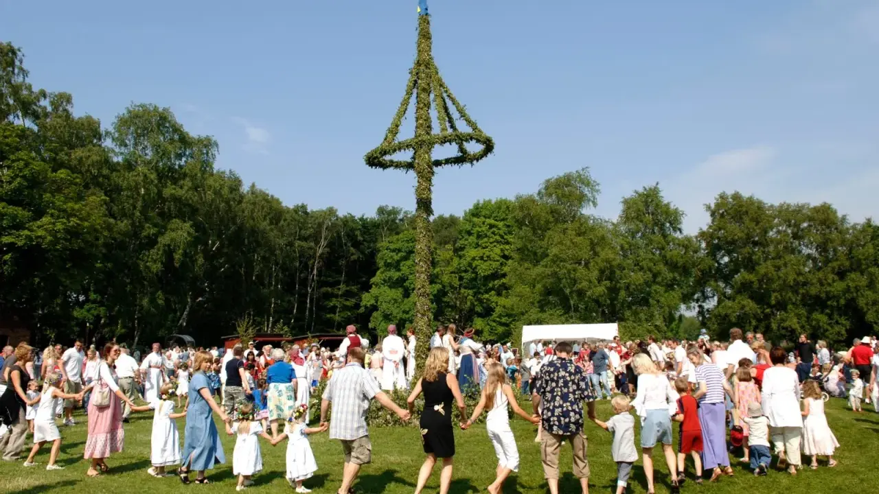Människor dansar runt en midsommarstång i en grönskande park. Många barn med blomsterkransar i håret deltar i firandet.