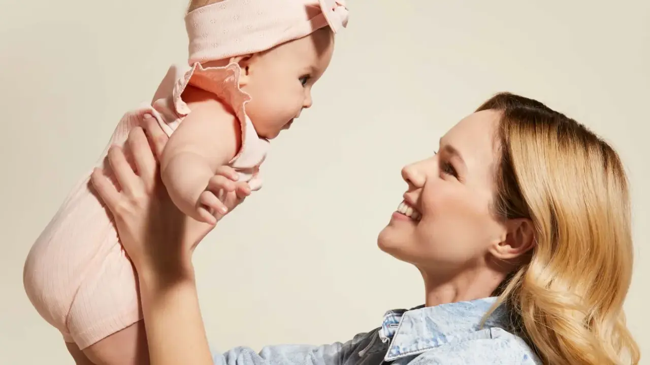 Mama podnosi dziecko w różowym body i opasce. Idealny przykład, jak ubierać noworodka latem – lekko i wygodnie.