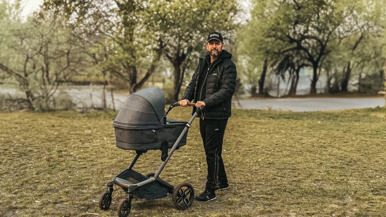 Mężczyzna w czapce z daszkiem i kurtce pcha nowoczesny wózek dla dziecka po trawie.