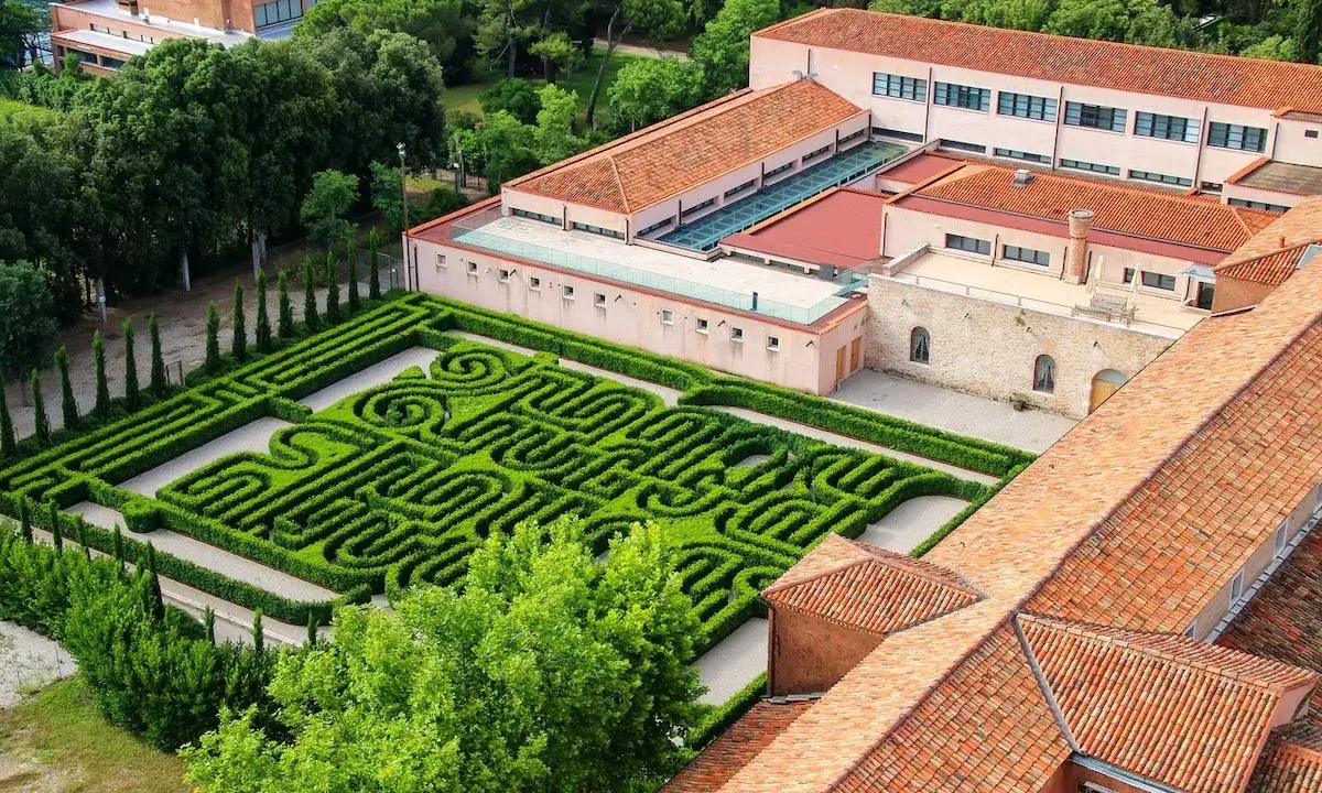 Un giardino con un intricato labirinto borges, circondato da edifici con tetti rossi e alberi verdi.