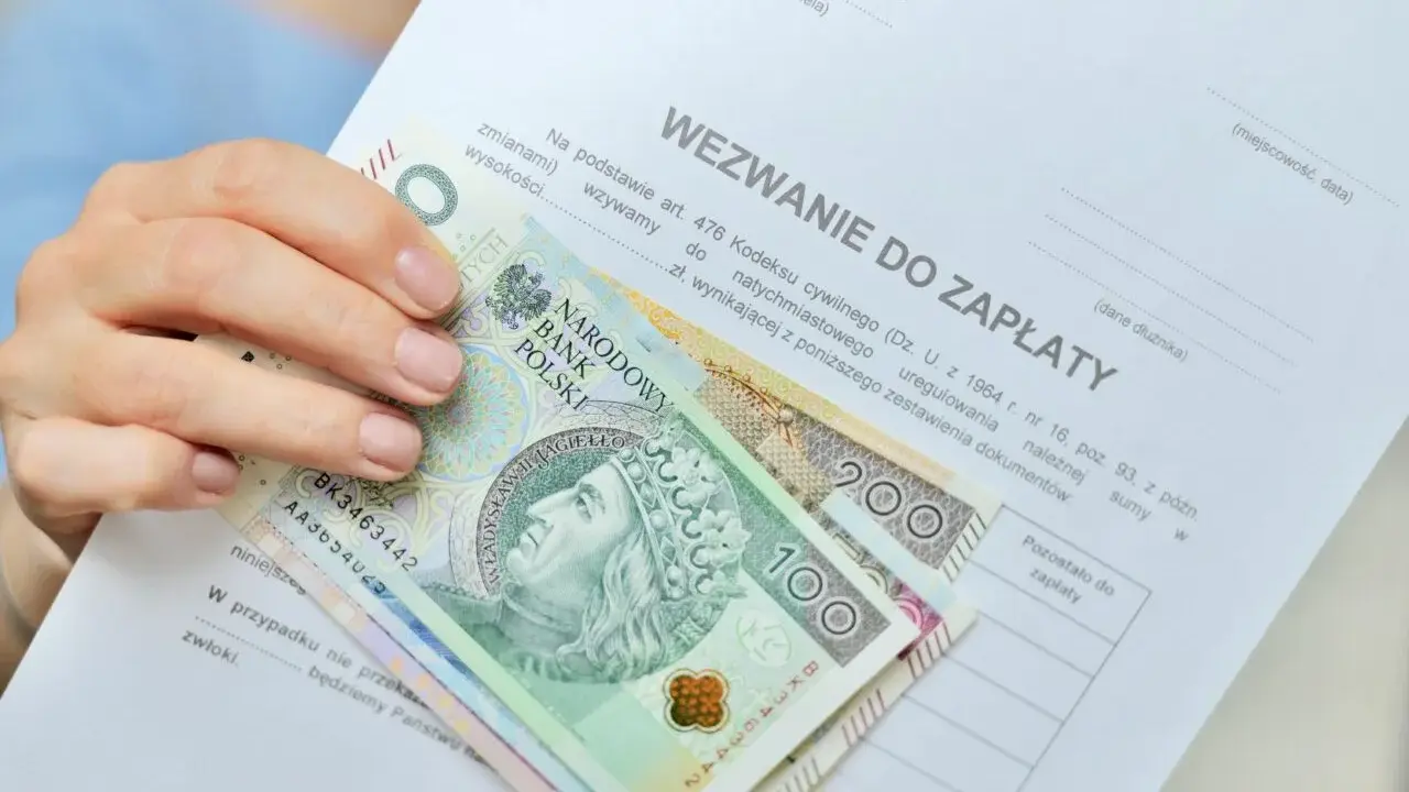Dłoń trzyma wezwanie do zapłaty i polskie banknoty 100 i 200 zł. Dokument zawiera informację o zawiadomieniu o uchyleniu zajęcia.