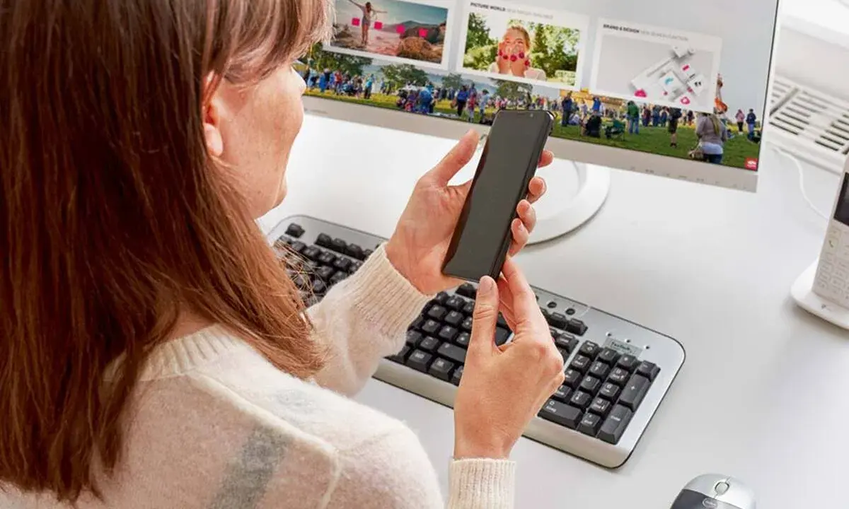 Kobieta trzyma smartfon przed monitorem komputera z klawiaturą i myszką.