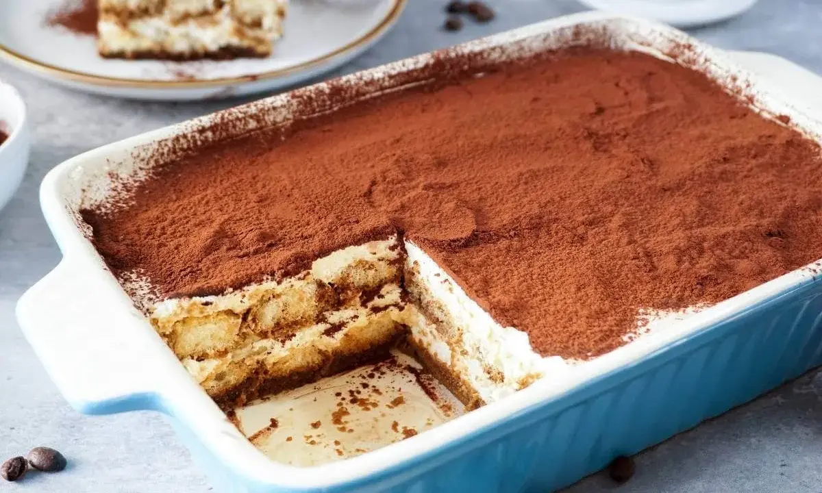 Ein köstliches Tiramisu ohne Ei, in einer blauen Auflaufform, bestäubt mit Kakao. Ein Stück ist bereits entnommen.