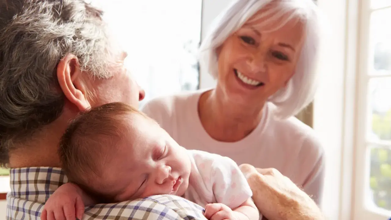 Los abuelos paternos sonríen mientras el abuelo sostiene al bebé dormido en su hombro.