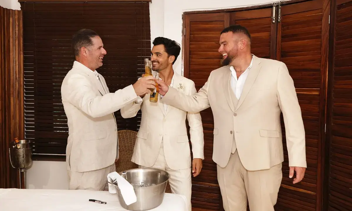 Tres hombres en trajes claros brindan con cerveza. El novio, en el centro, sonríe. Un momento de alegría antes del discurso de boda hermano.