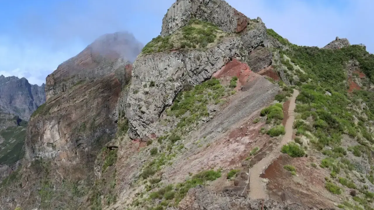 Steiler Bergpfad auf Madeira, ideal für die Packliste. Felsige Gipfel, grüne Vegetation und ein Weg, der sich durch die zerklüftete Landschaft schlängelt.