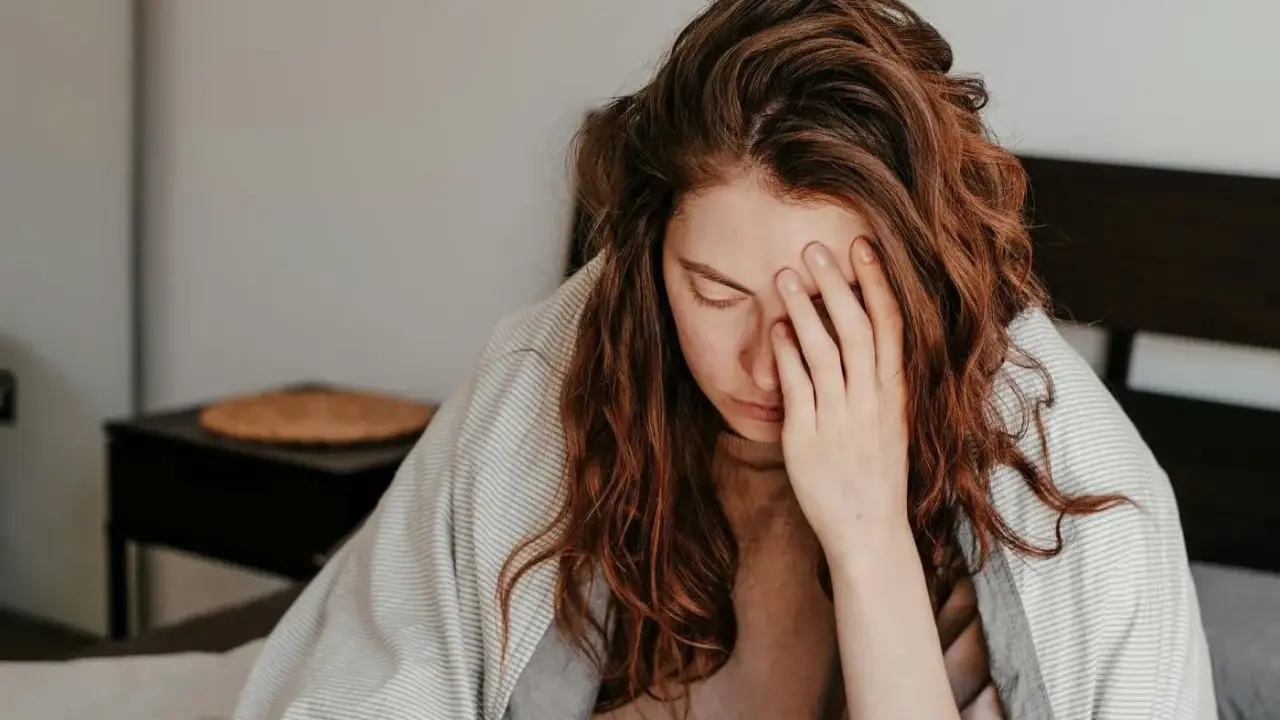 Kvinna med rött hår i sängen, täckt av filt, håller handen för ansiktet. Hon ser ut att ha feber, ett kolera symptom.