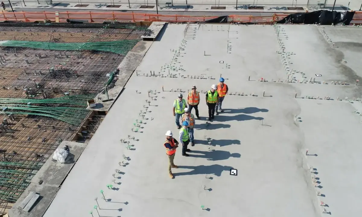 Grupa budowlańców na placu budowy, gdzie trwa wylewanie betonu. Zastanawiają się, ile kosztuje gruszka betonu, analizując postęp prac.
