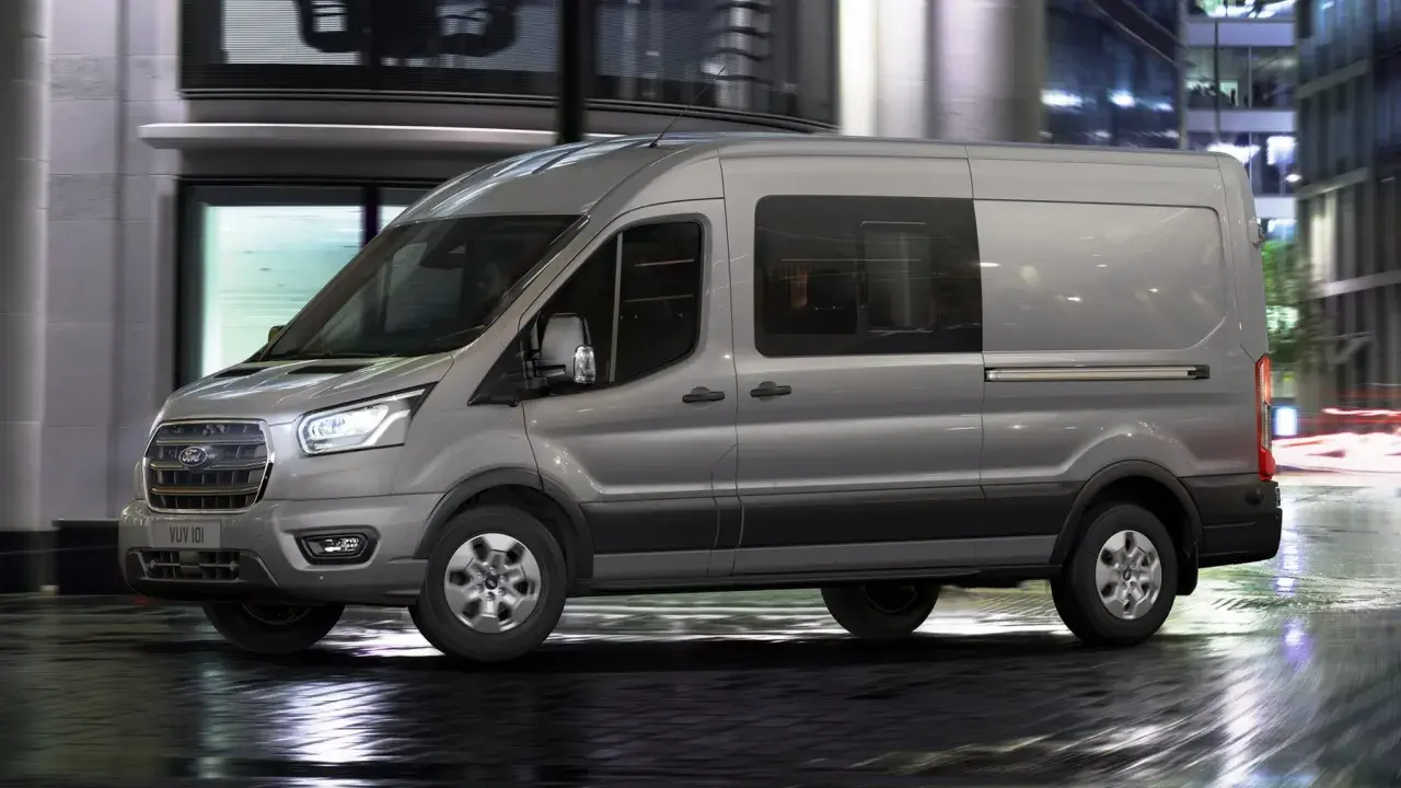 Camión N1 gris Ford Transit circulando por una calle urbana mojada de noche, con luces de edificios y tráfico difuminadas al fondo.