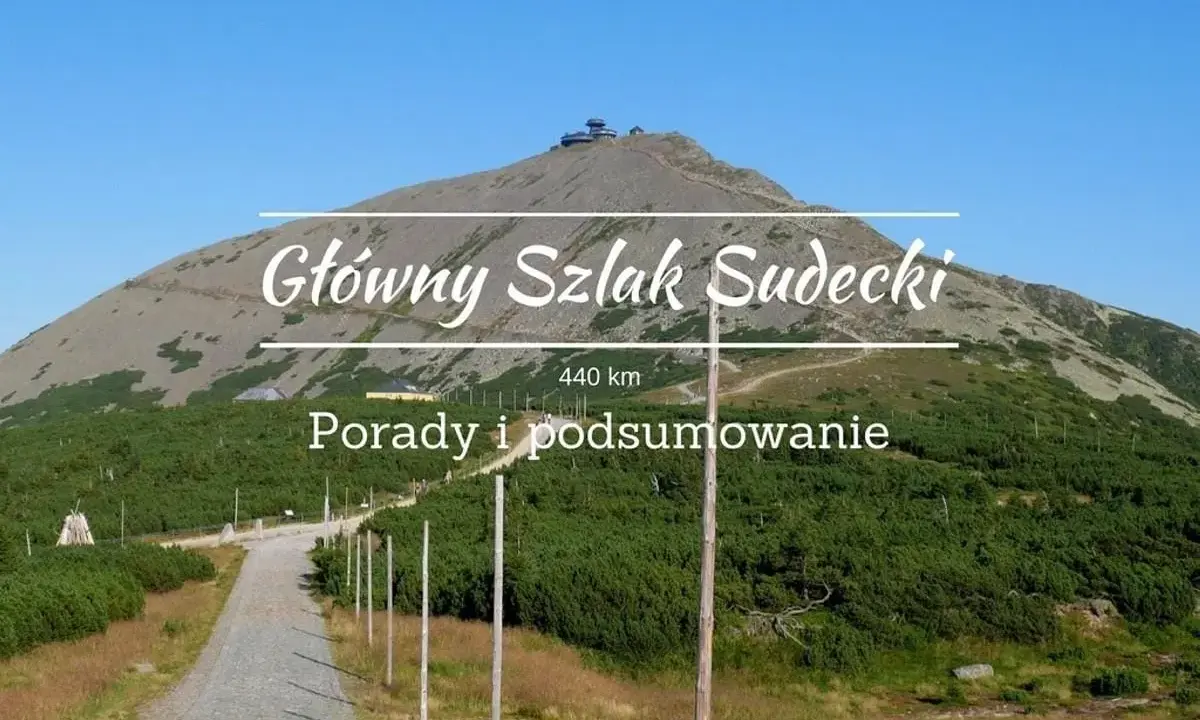Kamienista ścieżka prowadzi na szczyt góry, gdzie widać budynek. Nad krajobrazem widnieje napis "Główny Szlak Sudecki".