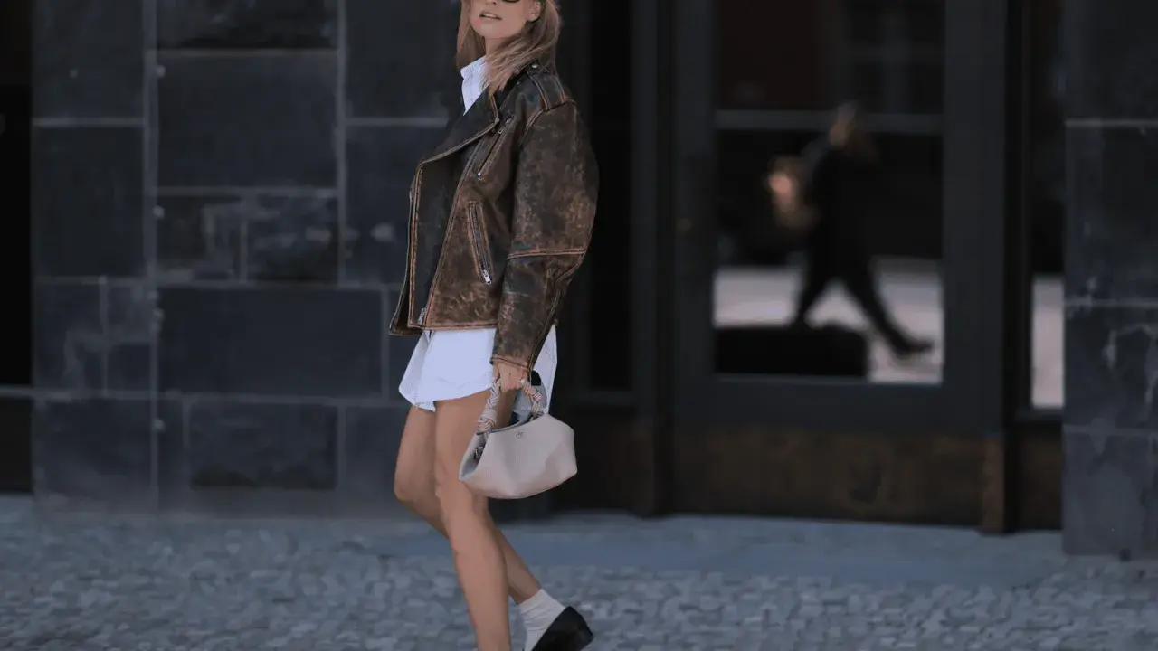 Stylish woman in a white dress, brown leather jacket, and black loafers with white socks walks confidently.