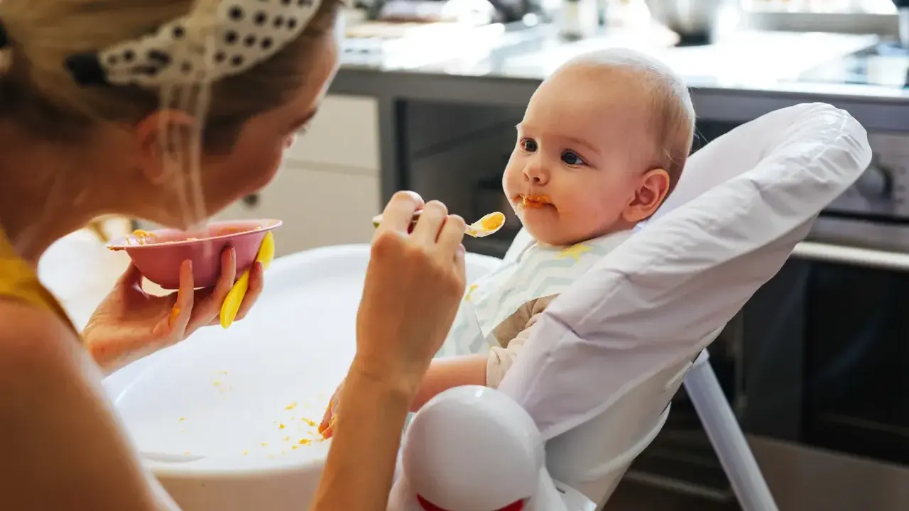 Mama karmi 7 miesięczne dziecko puree z marchewki. Maluch ma pomarańczowe ślady na buzi i śliniaczku.