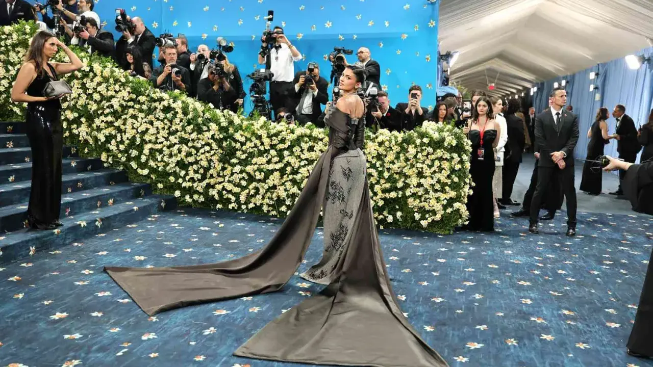 Kylie Jenner arrives at the Met Gala in a dramatic gown, posing for photographers amidst a sea of flowers and a blue, star-studded backdrop.