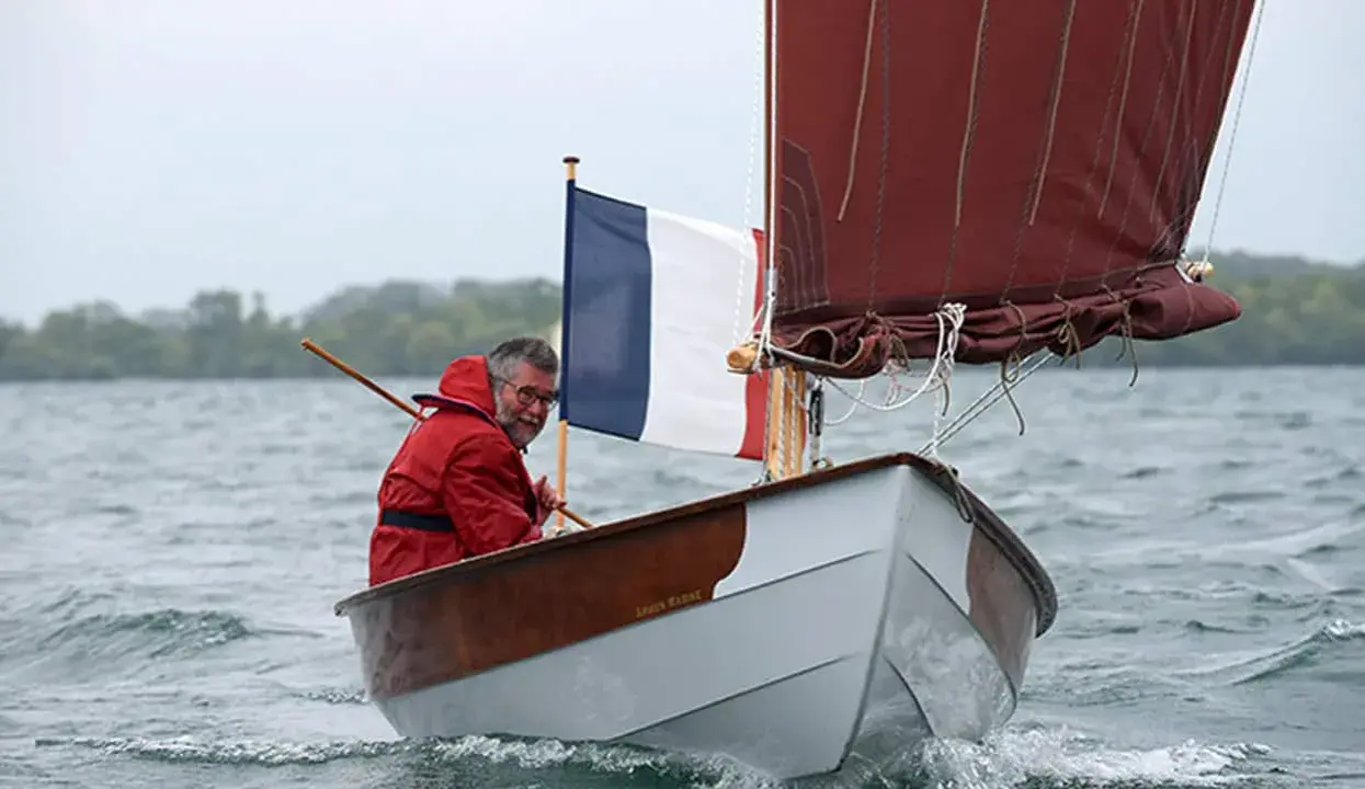Un homme en ciré rouge navigue sur son bateau, fier du résultat de son travail pour construire son bateau. Le drapeau français flotte fièrement.