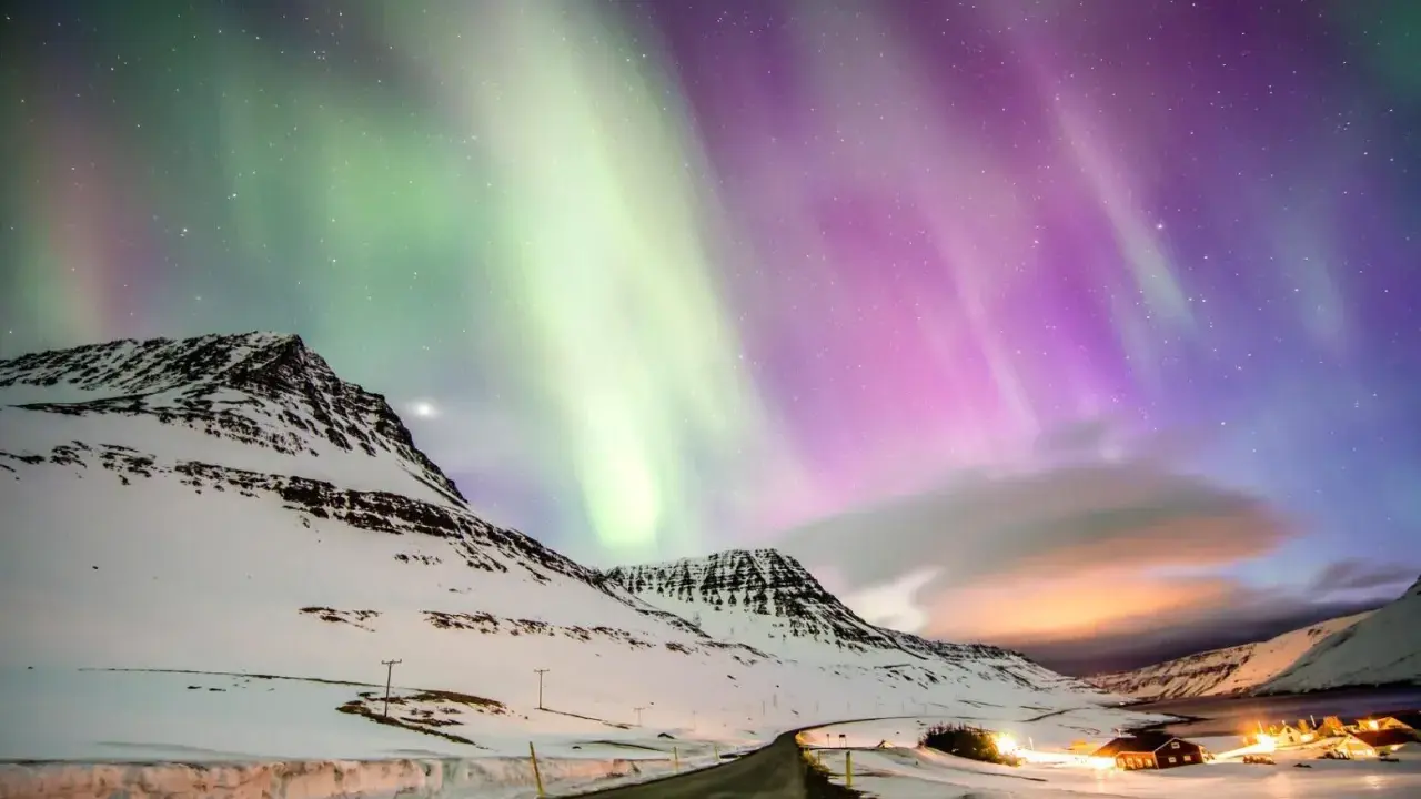 Abisko norrsken målar himlen i grönt och lila över snötäckta berg och en väg som leder mot upplysta hus.