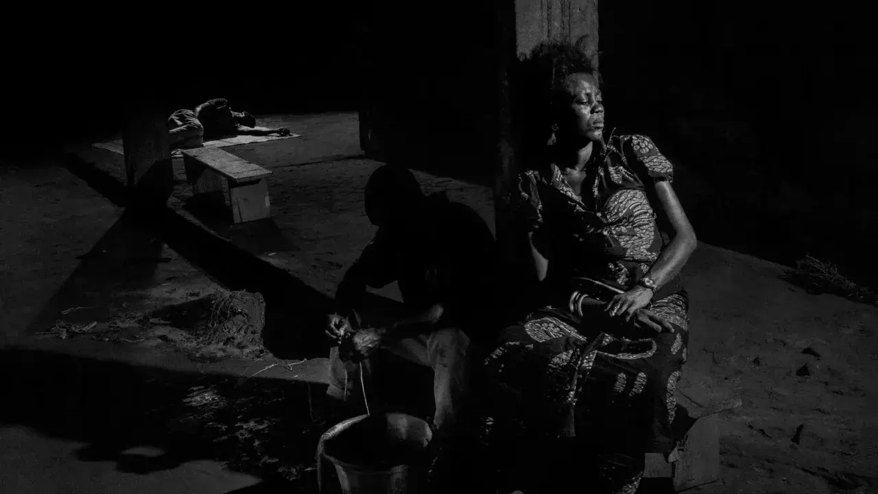 A woman in a patterned dress sits in shadow, eyes closed, while a man works with a bucket in the foreground. Alex Majoli captures a moment of quiet resilience.