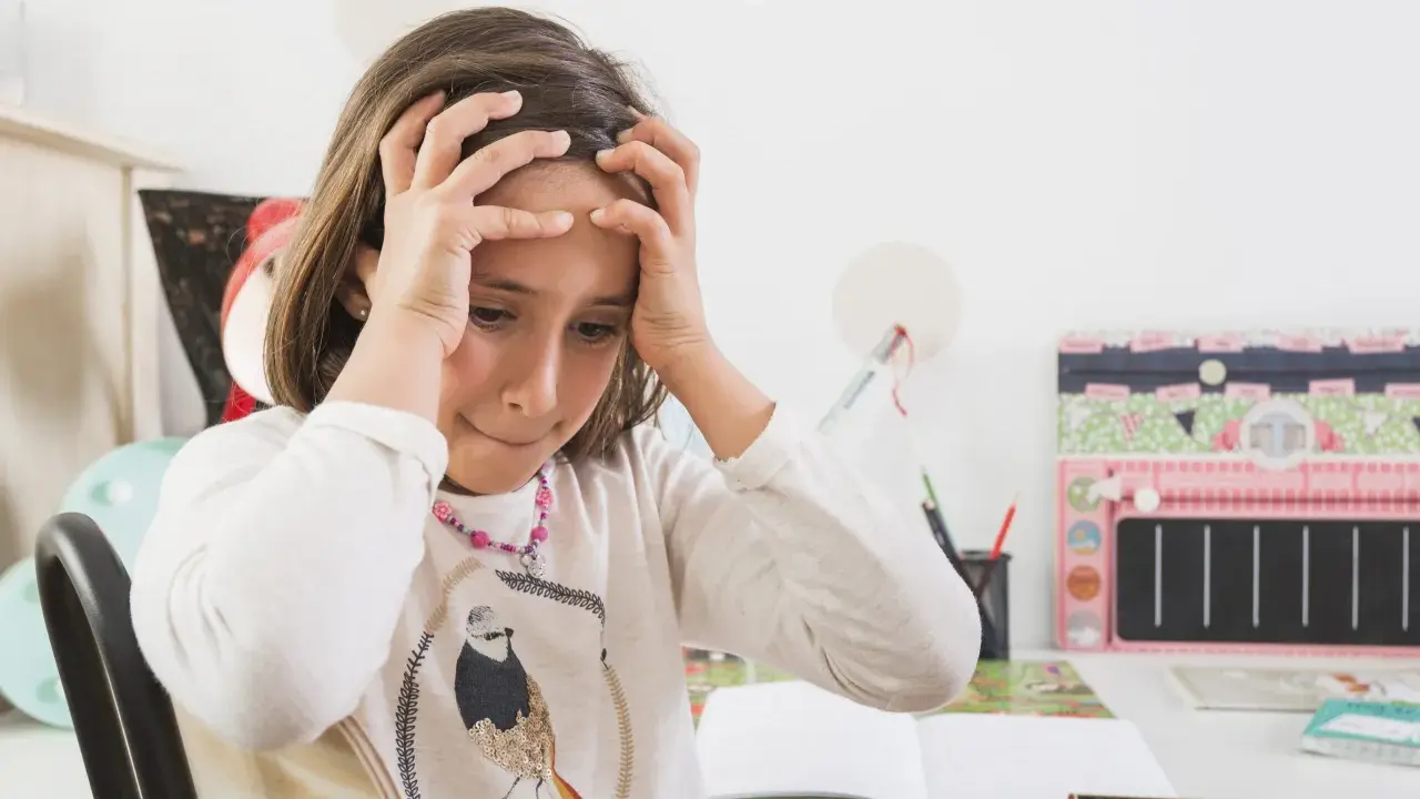 Dziewczynka z rękami na głowie, zmartwiona nauką. Jak rozładować stres u dziecka? Może przerwa na zabawę?