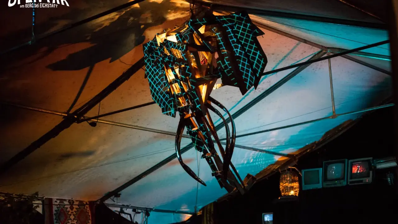 Abstrakte Skulptur mit blauem Gittermuster unter einem Zeltdach, beleuchtet von warmem und kühlem Licht.