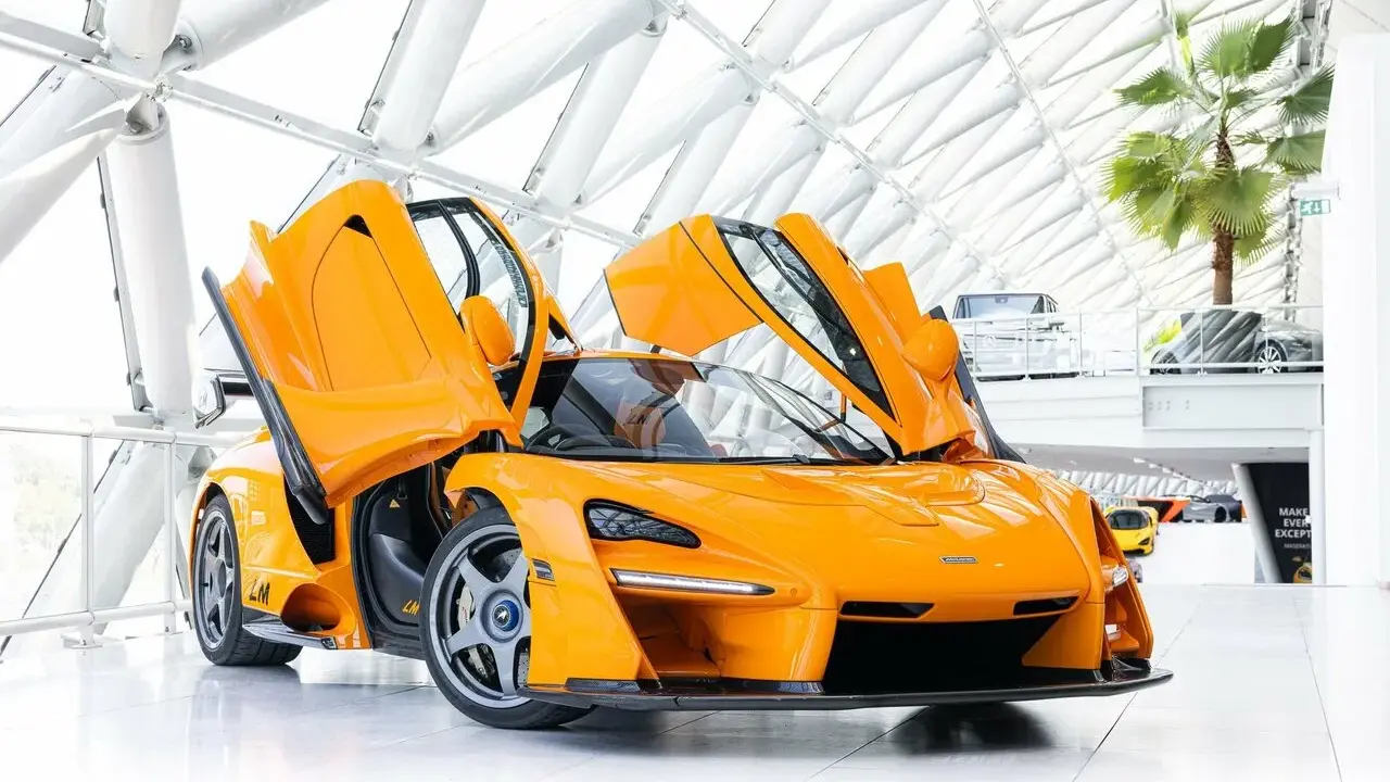 Ein leuchtend oranger McLaren Senna LM mit geöffneten Flügeltüren steht in einem modernen Gebäude mit Glasdach.