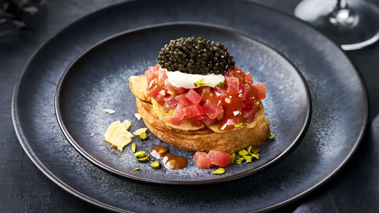 Kaviar Rezepte Vorspeise: Thunfisch-Tatar auf geröstetem Brot, gekrönt mit Kaviar und einem Klecks Crème fraîche. Pistazien und Chips garnieren den Teller.