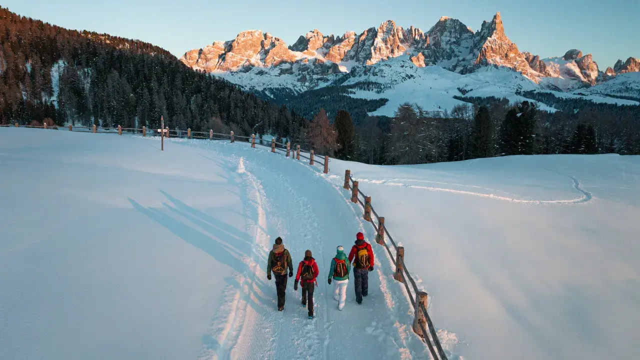 Quattro escursionisti camminano su un sentiero innevato al Passo di Lavazè, con le maestose vette dolomitiche illuminate dal sole al tramonto.