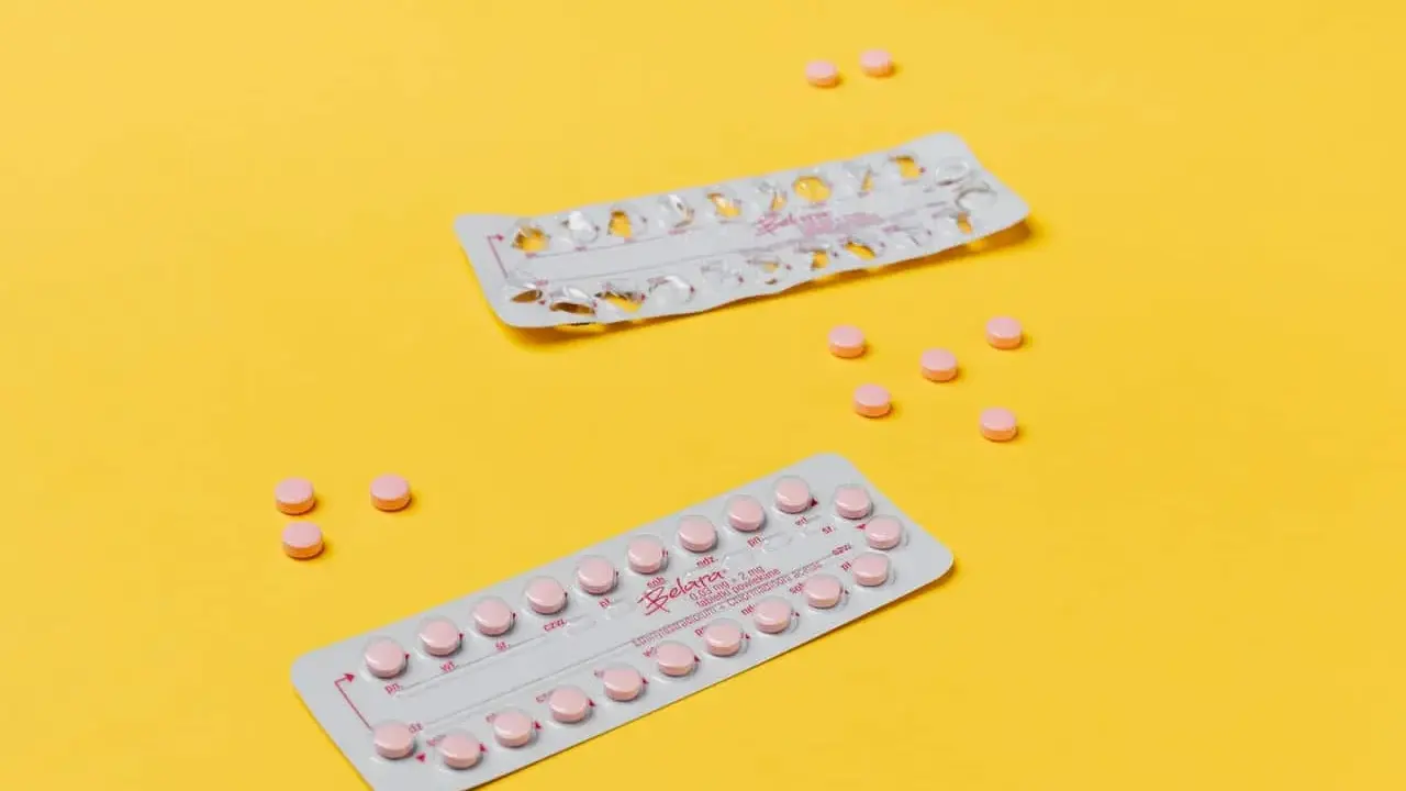 Dos blísteres de pastillas anticonceptivas y varias pastillas sueltas, rosas, sobre fondo amarillo. Ayudan a regular la regla.