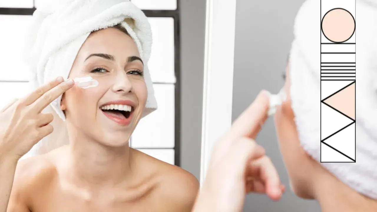 Donna sorridente con asciugamano in testa applica crema sul viso, parte della sua routine skincare.