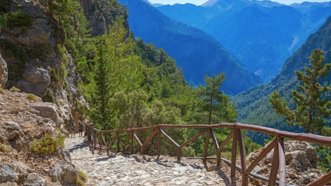 Wanderweg in einer atemberaubenden **Schlucht auf Kreta**. Steile Felswände, grüne Bäume und ein tiefer Abgrund, gesichert durch einen hölzernen Zaun.