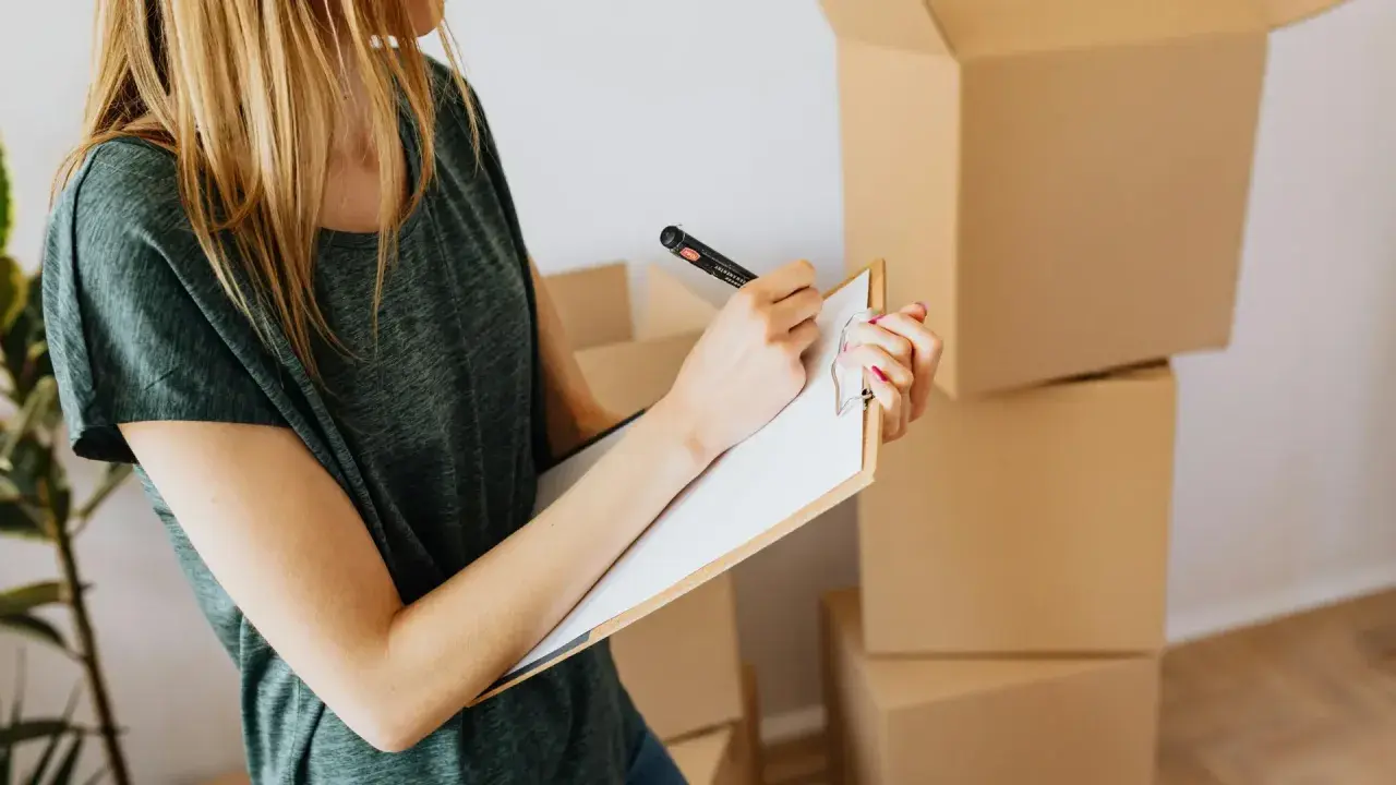 Mujer rubia con camiseta verde, revisando una lista en un portapapeles, rodeada de cajas de mudanza. El arte 19 lau inspira la organización.