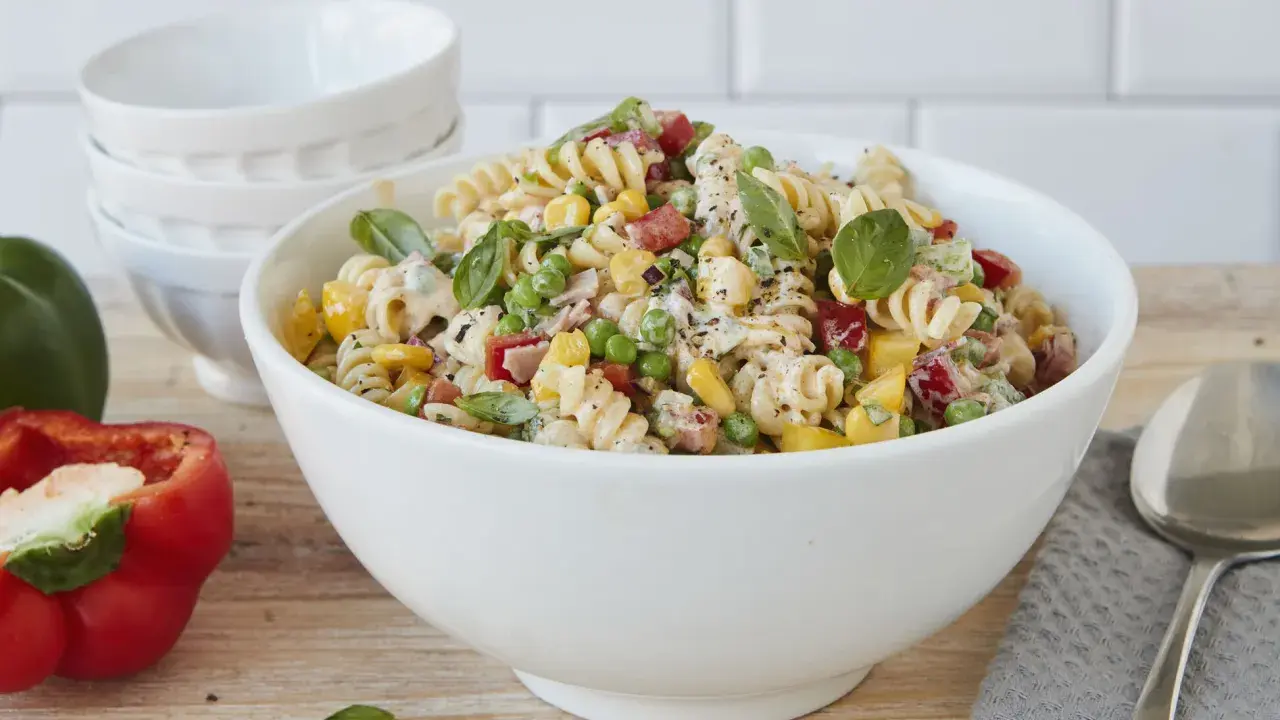 Ein bunter Nudelsalat ohne Mayonnaise mit Spiralnudeln, Erbsen, Mais, Paprika und frischem Basilikum in einer weißen Schüssel.