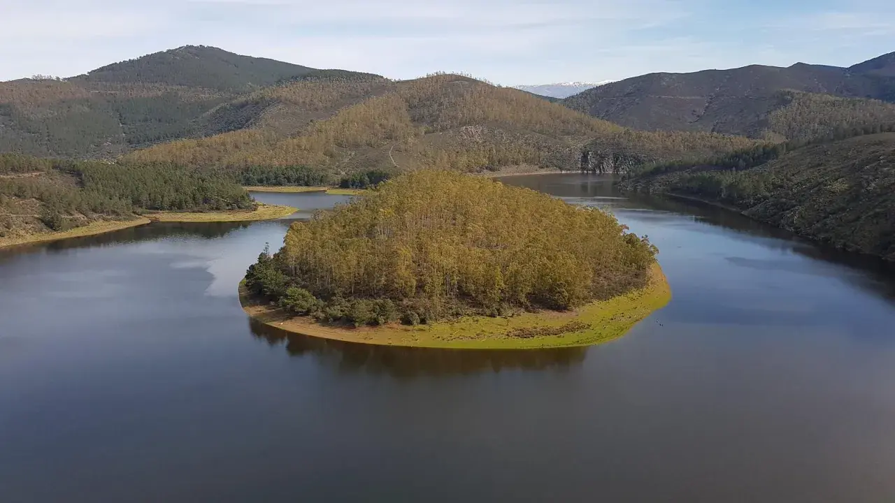 El meandro del Melero, una isla verde rodeada de agua serena, se abre paso entre colinas boscosas bajo un cielo azul.