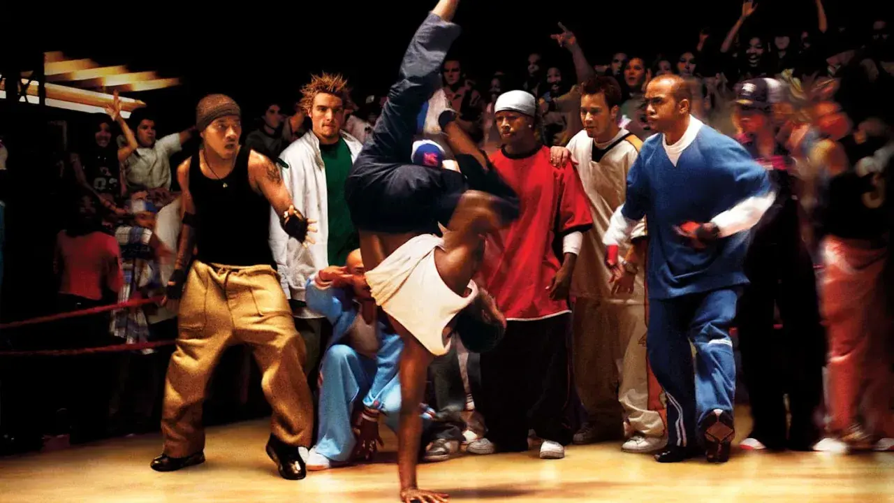 Energetyczna scena z filmów o tańcu. Breakdancer wykonuje akrobację, otoczony przez widzów i innych tancerzy.