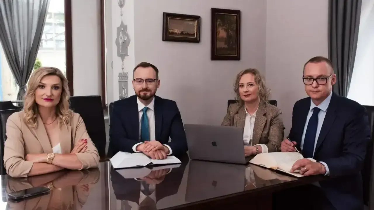 Cztery osoby w strojach biznesowych siedzą przy stole, przed nimi dokumenty i laptop.