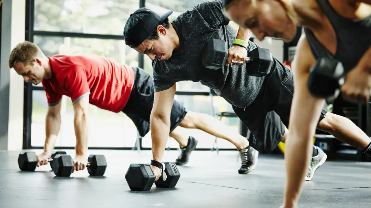 Drei Personen machen ein intensives Crossfit Training mit Kurzhanteln. Sie sind in einer Plank-Position und ziehen abwechselnd eine Hantel hoch.