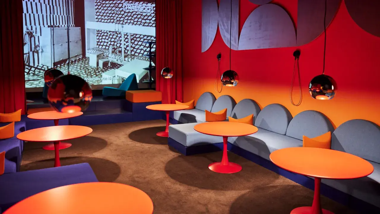 En färgstark lounge med soffor, orange bord och en projektion av Nanna Ditzels design.